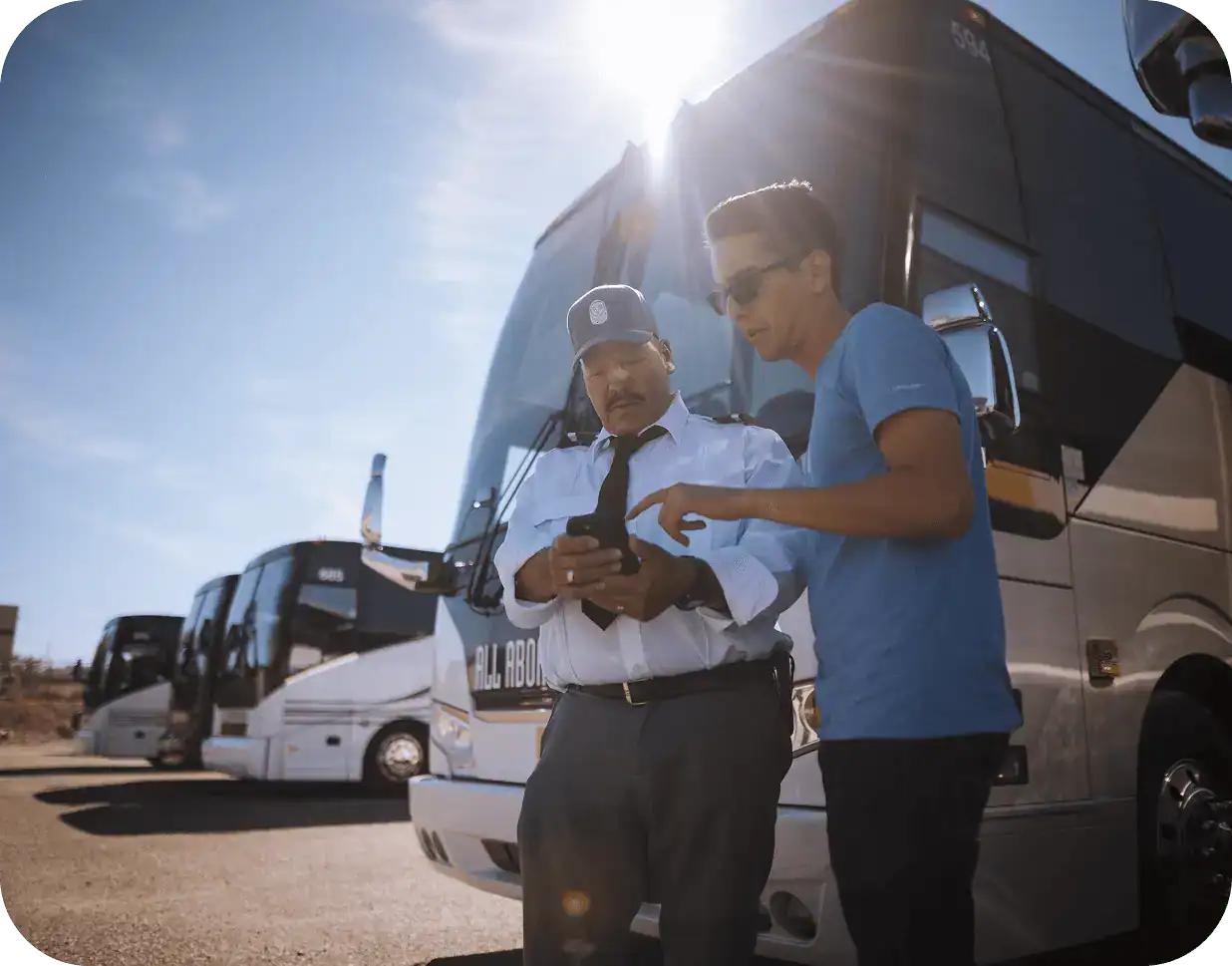 An All Aboard America bus driver and customer stand together in front of several buses while looking at a cell phone to review trip details.