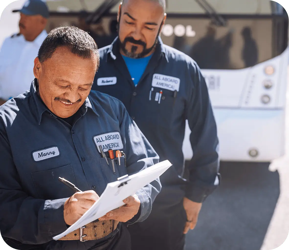 Two bus mechanics stand together while one smiles and signs some paperwork.