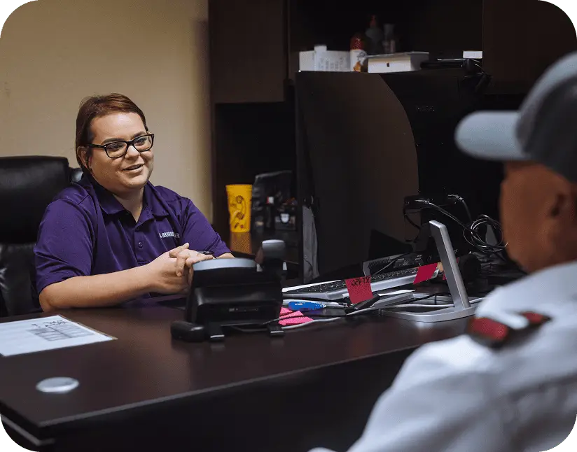 An office employee sits at a desk smiling and talking with a bus driver sitting opposite.