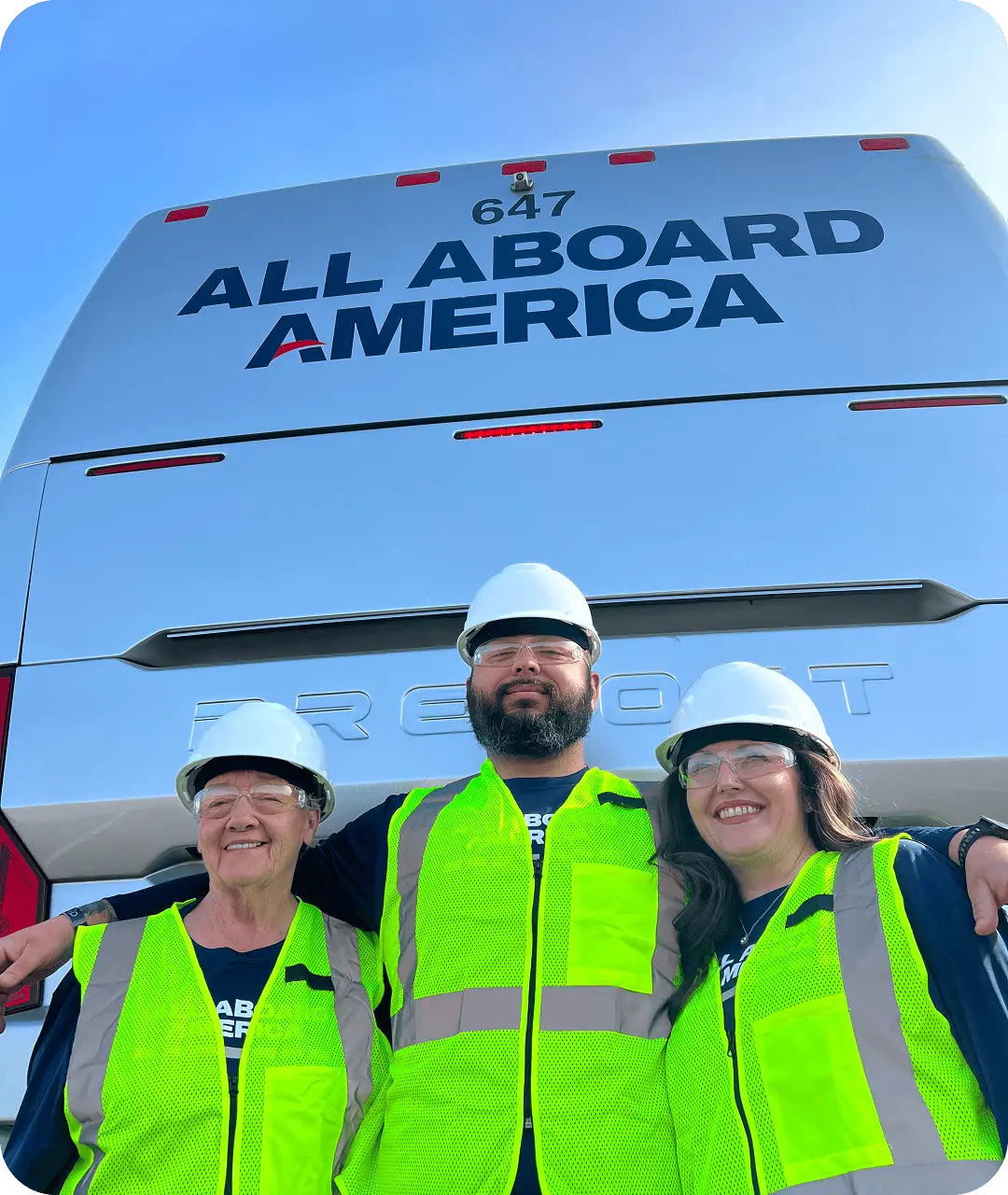 3 All Aboard America employees, wearing hard hats and green safety vests, are standing behind an All Aboard America bus
