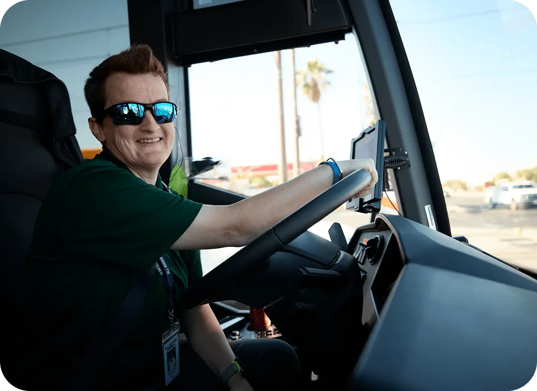 A smiling All Aboard America driver wearing sunglasses sits behind the wheel of a motorcoach