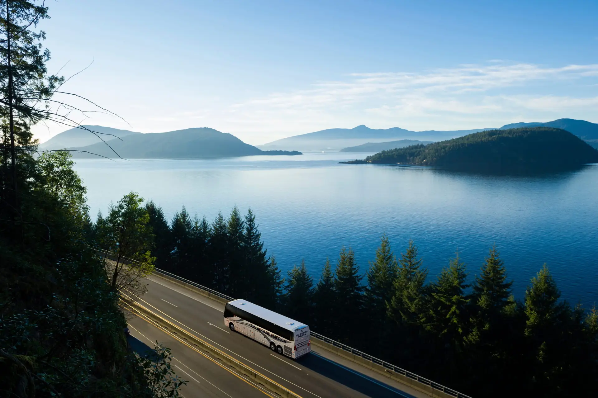 A motorcoach travels along a scenic coastal highway with mountains and a lake in the background