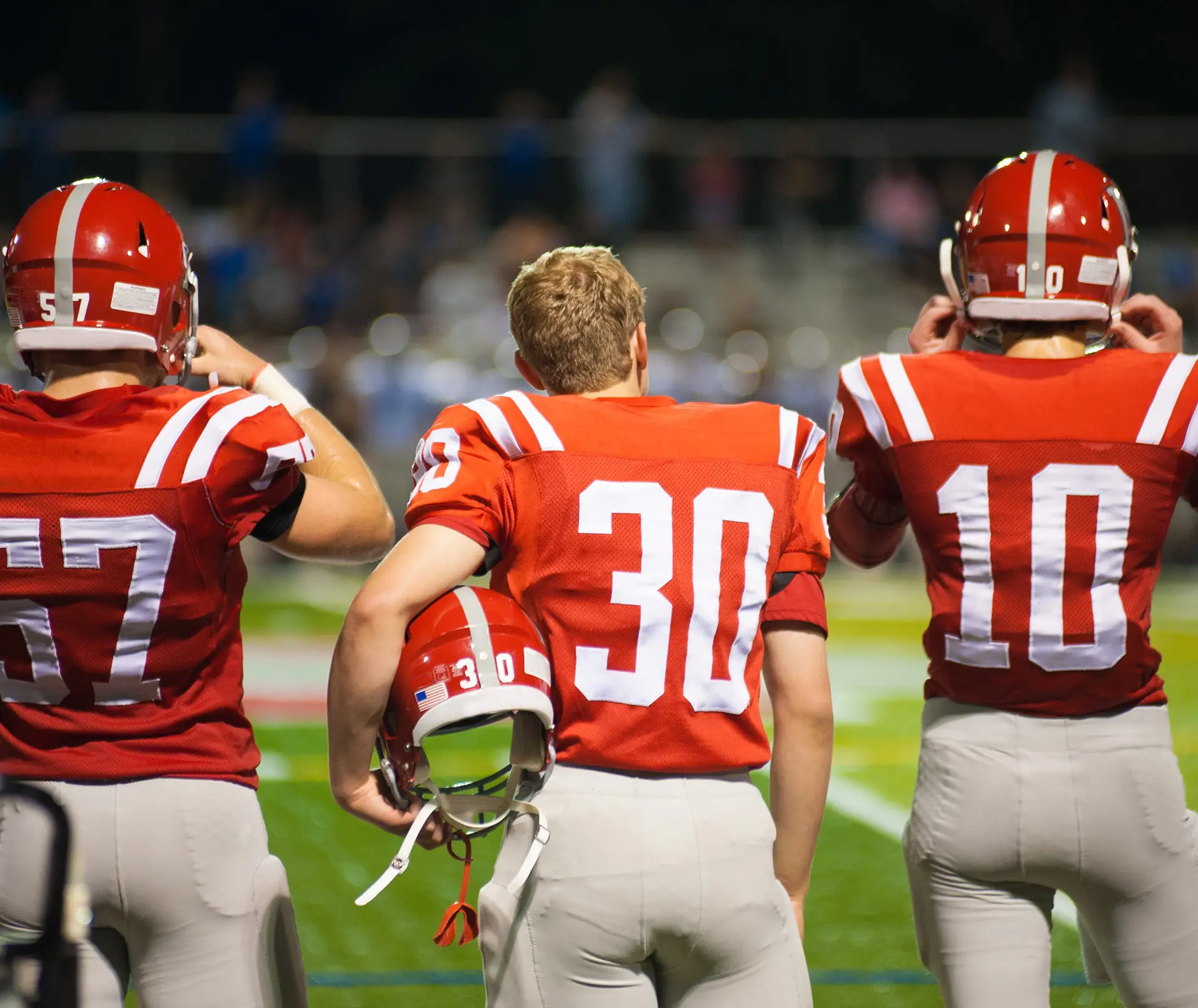 High school football players in red jerseys stand on the sideline during a night game