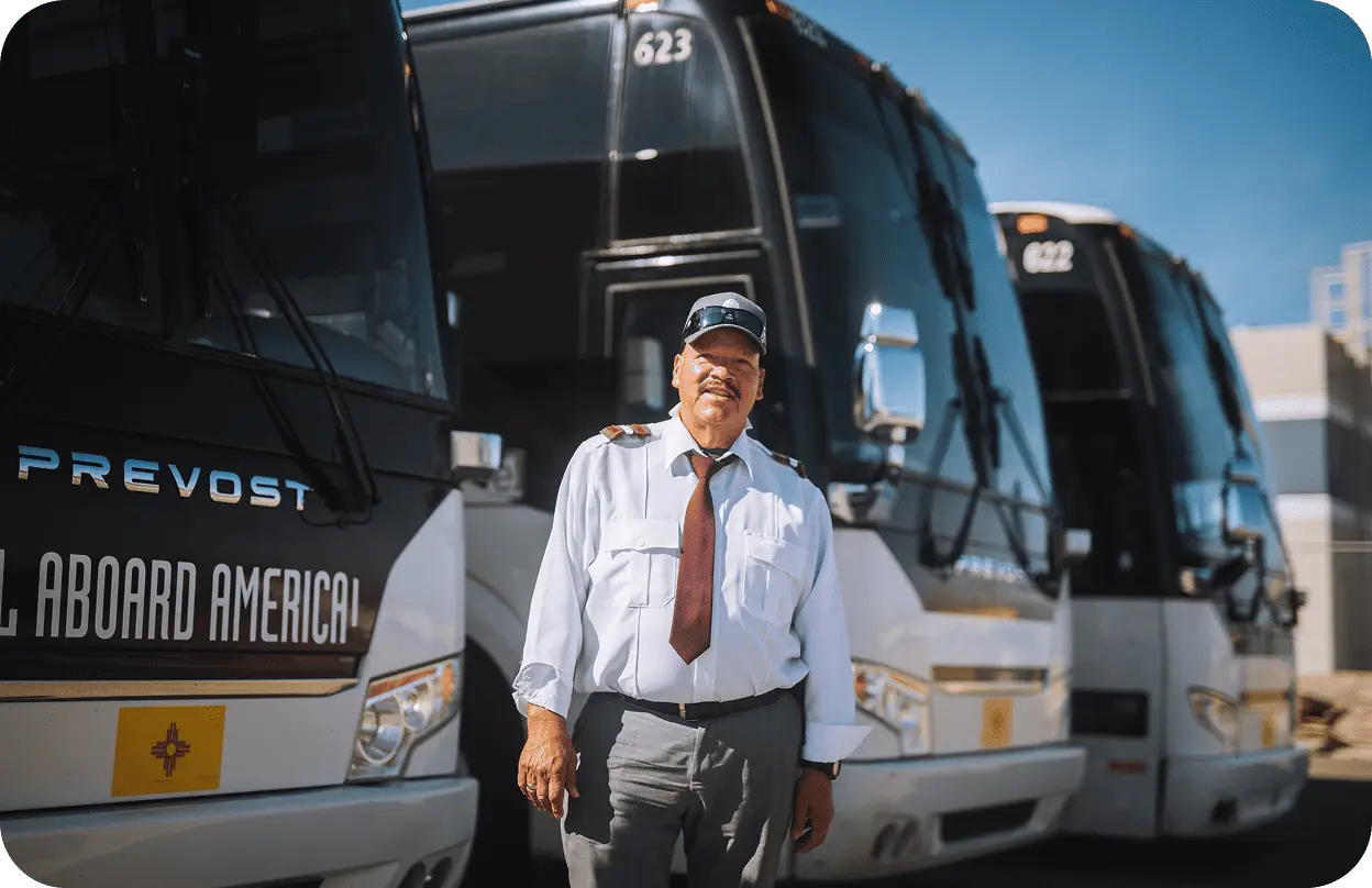An All Aboard America driver in uniform and cap stands proudly in front of a row of Prevost motorcoaches