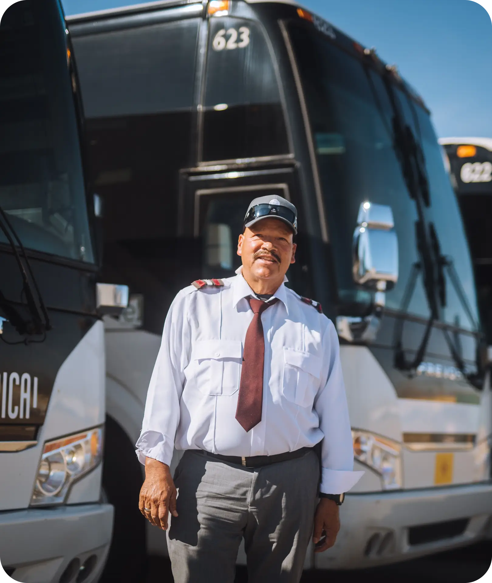 An All Aboard America driver in uniform and cap stands proudly in front of motorcoaches at the fleet depot