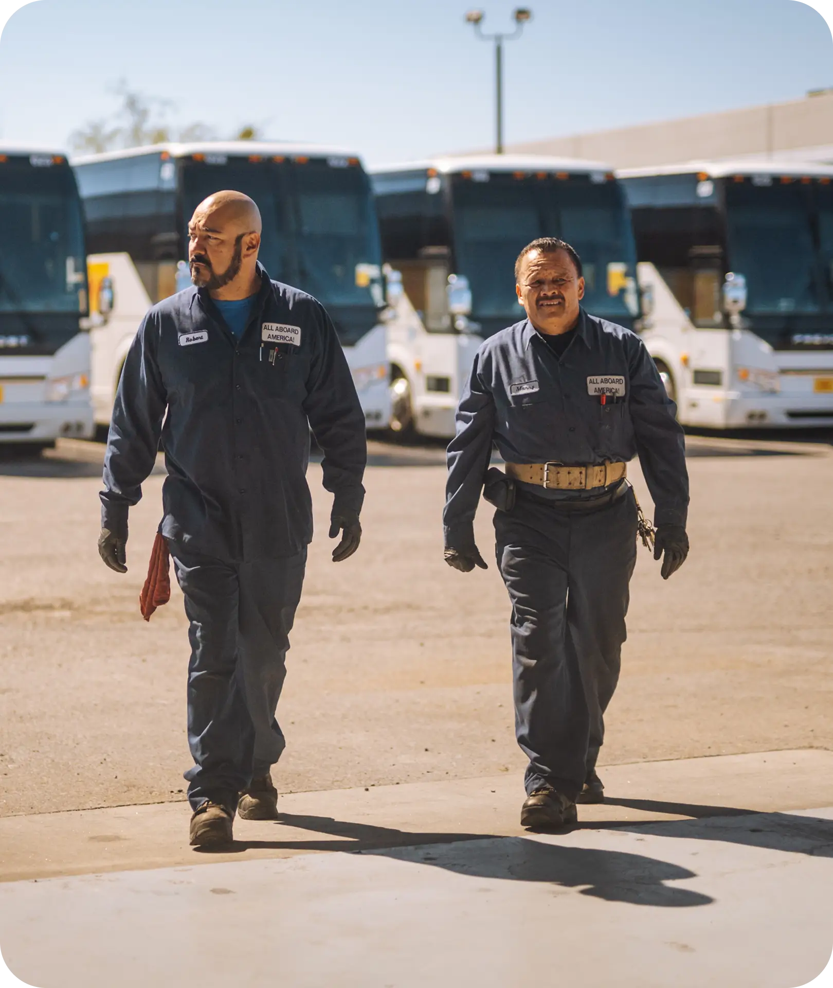 Two All Aboard America mechanics in uniform walk across a lot in front of a parked fleet of buses