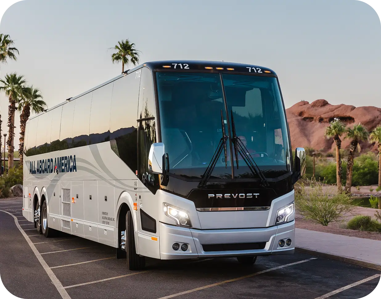 An All Aboard America Prevost motorcoach parked at dusk with palm trees and desert rock formations for industrial workforce transport