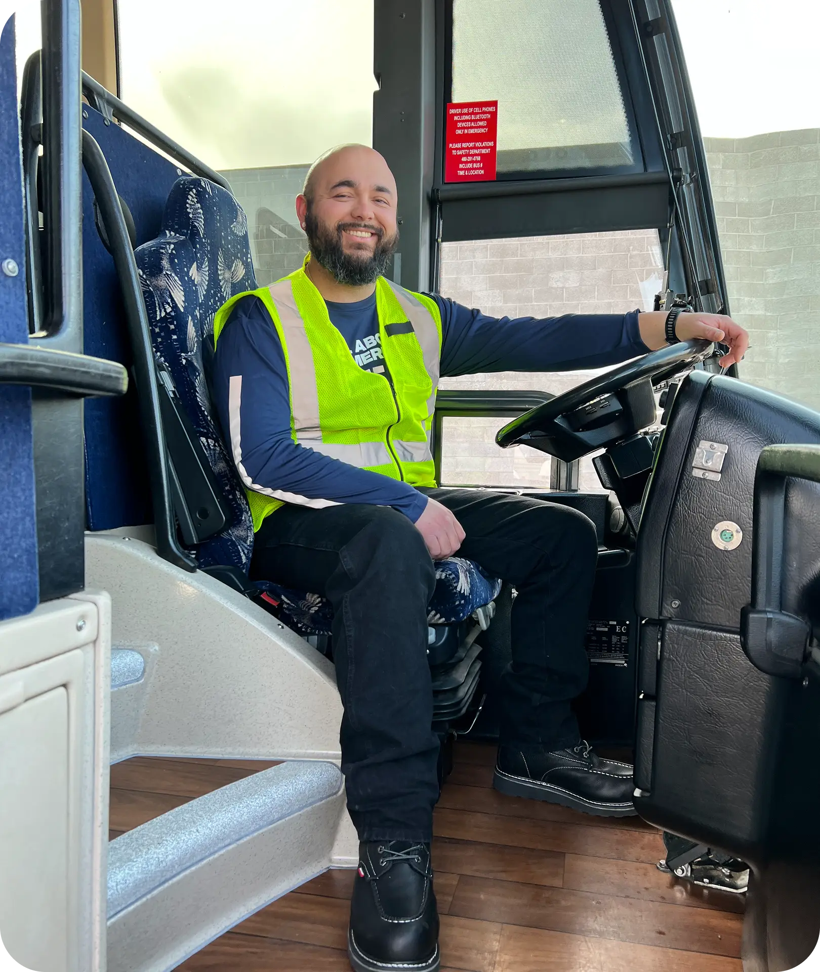 A smiling All Aboard America driver in a safety vest sits behind the wheel of an industrial workforce bus